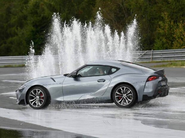 Ein grauer Toyota Supra ist seitlich zu sehen, er fährt auf durchnässter, spiegelnder Strecke auf eine breite, weiße Wasserfontäne zu. 