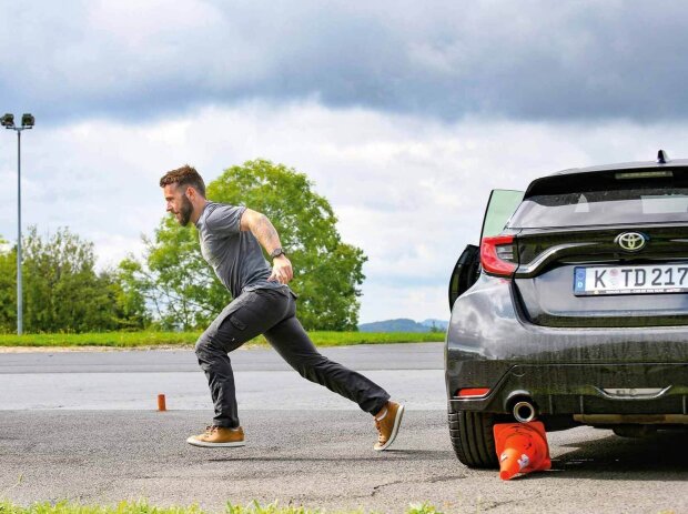 Ein Mann sprintet nach links von einem schwarzen Toyota Yaris weg, der von hinten zu sehen ist. 