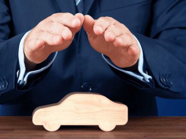 A man in a blue suit holds his hands protectively over a wooden car that is standing on a dark wooden table in front of him.