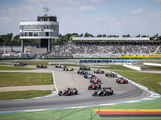 Mehrere Autos auf der Rennstrecke biegen in eine Rechtskurve ein. Im Hintergrund der Turm des Baden-Wurttemberg-Center und volle Tribünen des Hockenheimrings.