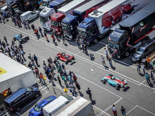 Von oben ein Blick auf eines Gasse des Fahrerlagers. Zwischen LKWs und Zelten der Teams fahren durch eine zuschauergesäumte Gasse drei Formel-Rennwagen