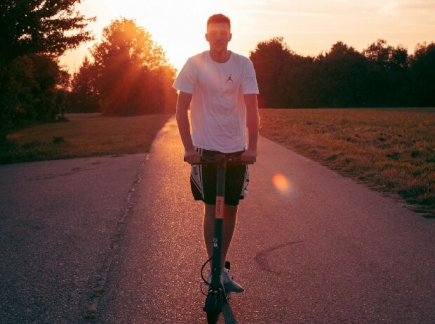 E-Scooter Versicherung: Junger Mann fährt E-Scooter auf einem Feldweg bei Sonnenuntergang E-Scooter Versicherung: Junger Mann fährt E-Scooter auf einem Feldweg bei Sonnenuntergang
